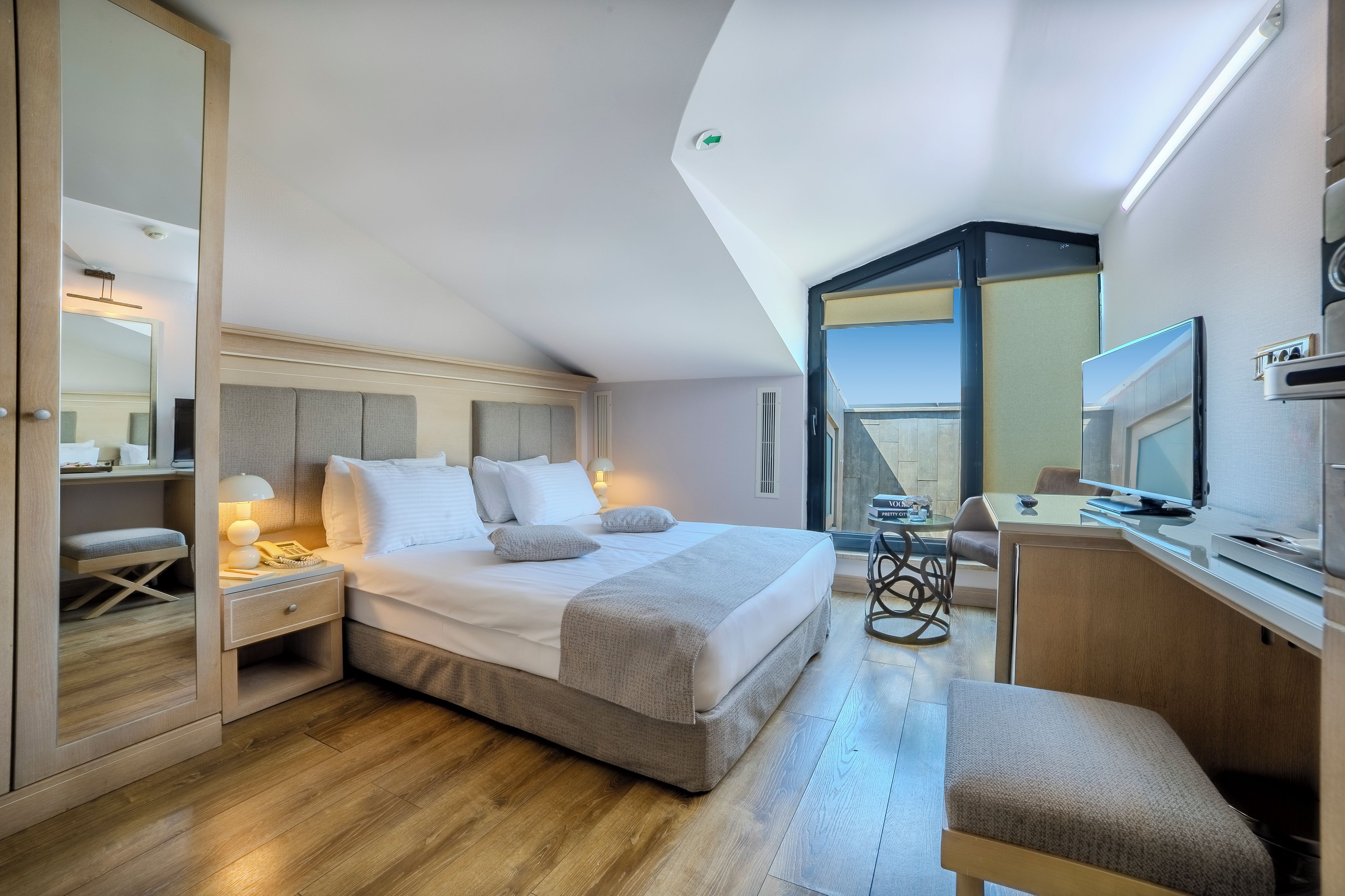 A modern hotel room with a large bed, a desk with a computer, a chair, and a television.