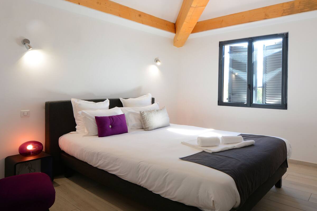 Una cama bien hecha con ropa de cama blanca y oscura, una almohada morada y una lámpara de noche.