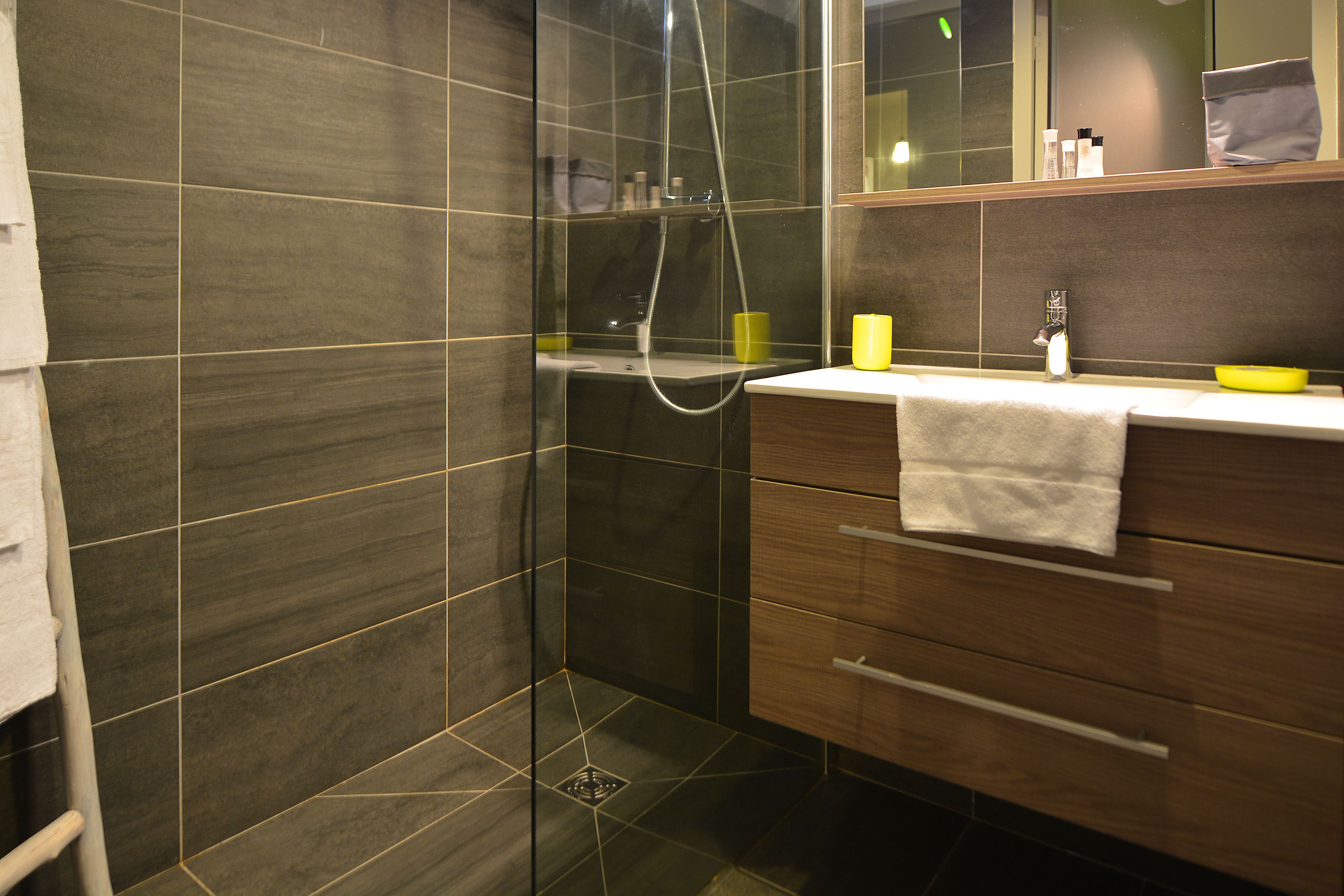 A modern bathroom with dark tiles, a glass shower enclosure, a wooden vanity with a yellow soap dish, and a mirror above the sink.