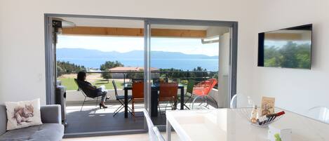 A modern living room with a sofa, a dining table, and a balcony with a view of the sea and mountains.