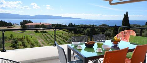 A balcony with a table set for two, chairs, and a view of the sea and mountains.