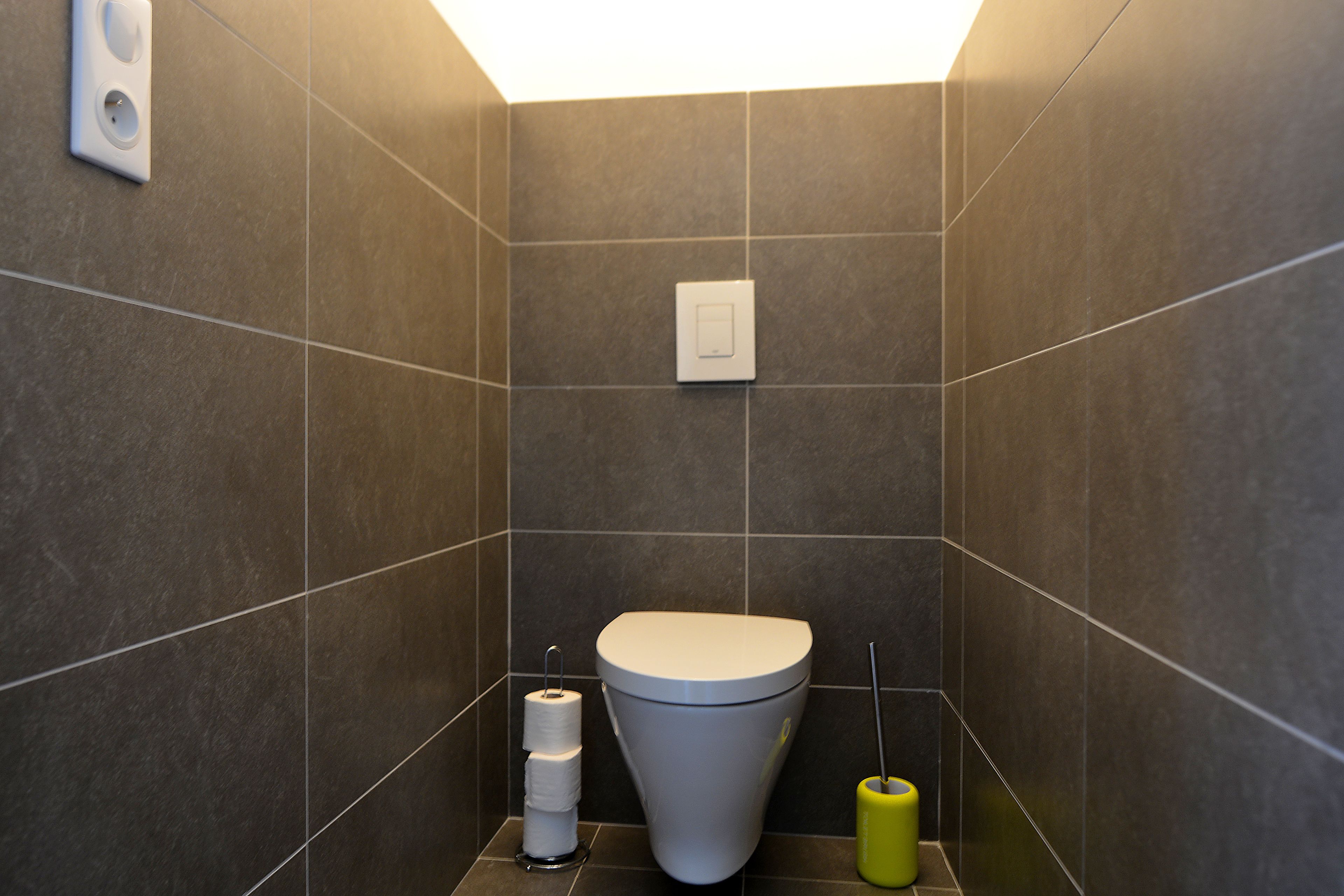 Une petite salle de bain moderne équipée de toilettes, d’un porte-rouleau de papier toilette et d’une brosse de nettoyage jaune.