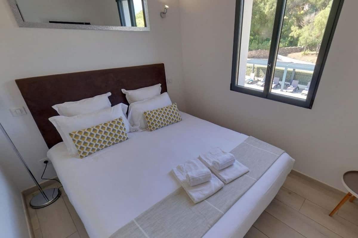 Una cama bien hecha con sábanas blancas y almohadones estampados, una mesita de noche con un control remoto y una pequeña planta, y una ventana con vista a un área de estar al aire libre.