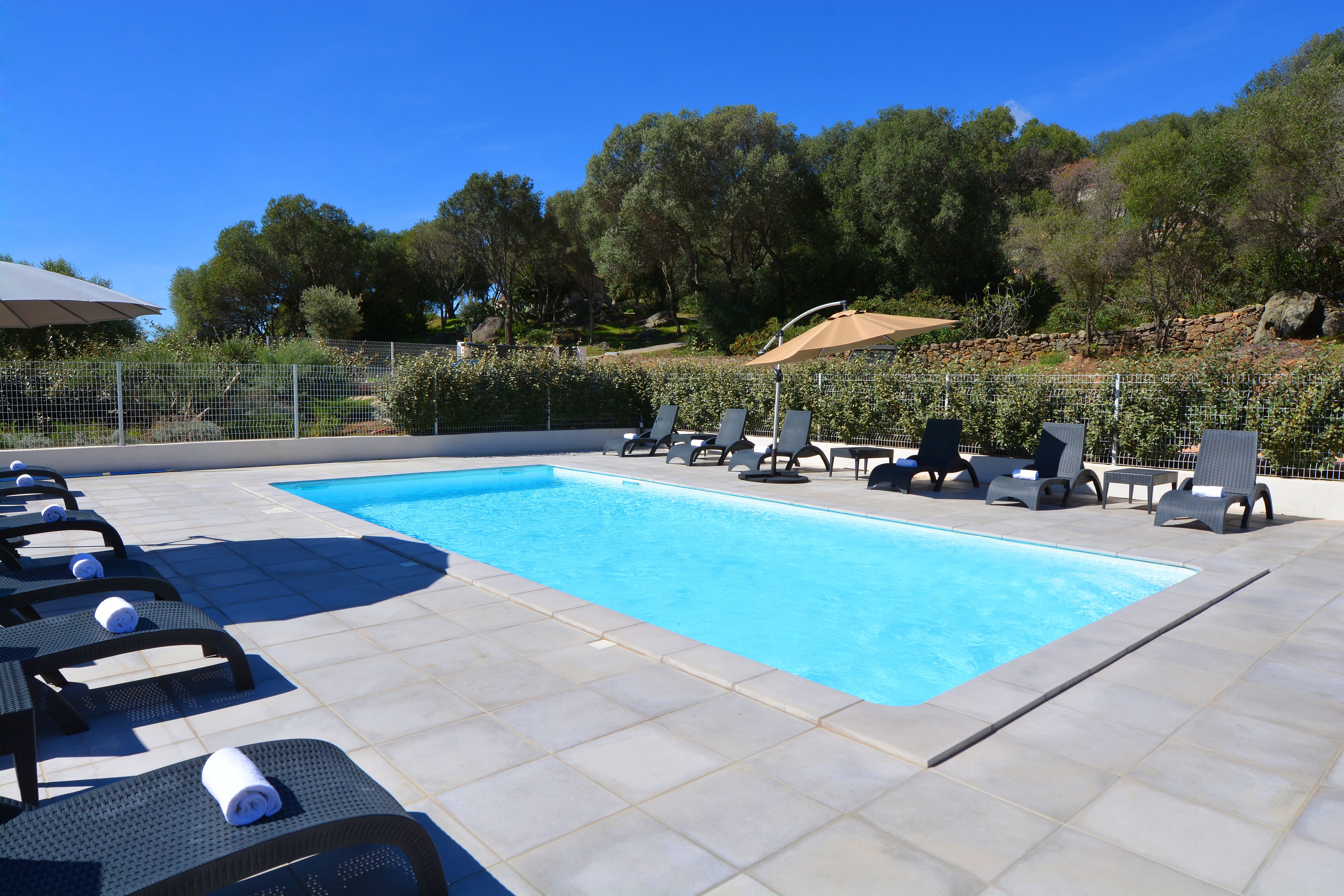 A swimming pool with lounge chairs and umbrellas, surrounded by a fence and trees.