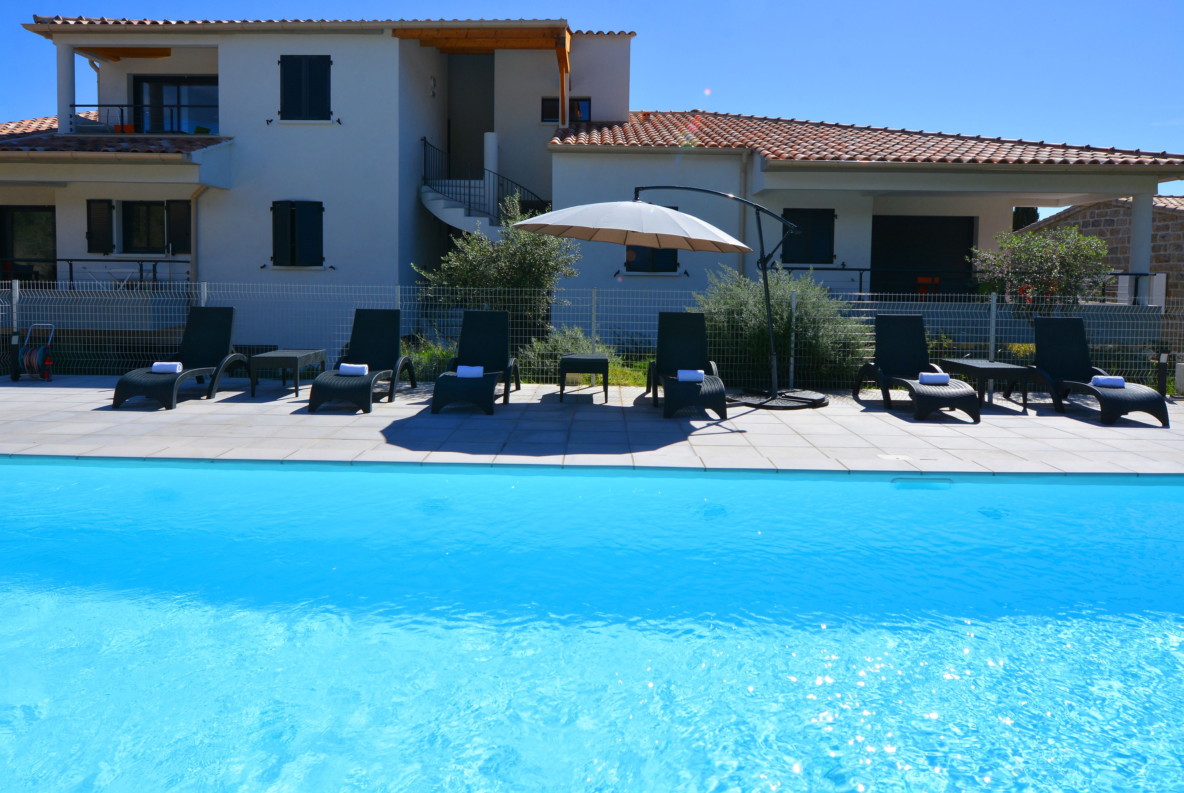 A pool area with lounge chairs and umbrellas, a clear blue pool, and a building with a tiled roof and balconies.
