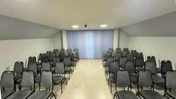 Konferenssitila, jossa on riveissä järjestettyjä tuoleja, jotka ovat vastakkain pienen esiintymislavan kanssa. Tilassa on kattovalaisin ja ikkuna, jossa on verhot.