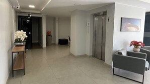 Un vestíbulo amplio y moderno con piso de baldosas, una recepción con un jarrón de flores, un área de descanso con una silla y una planta en maceta, y un ascensor.