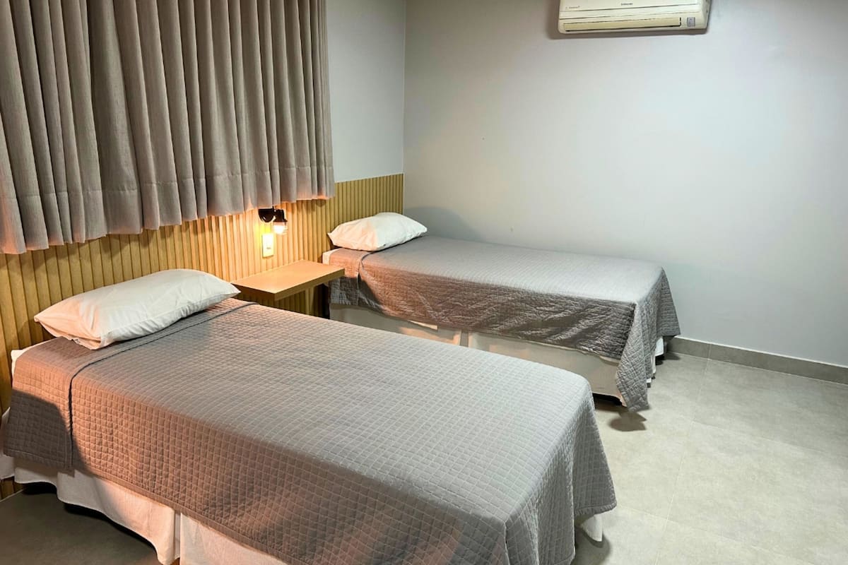 Dos camas individuales con ropa de cama gris, un cabecero de madera, una mesita pequeña y un aire acondicionado de pared.