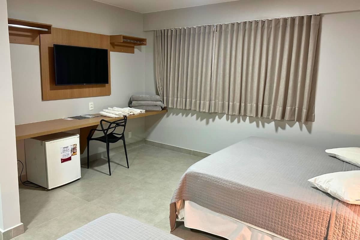Habitación de hotel con una cama, un escritorio con computadora, televisor montado en la pared y una ventana con cortinas.
