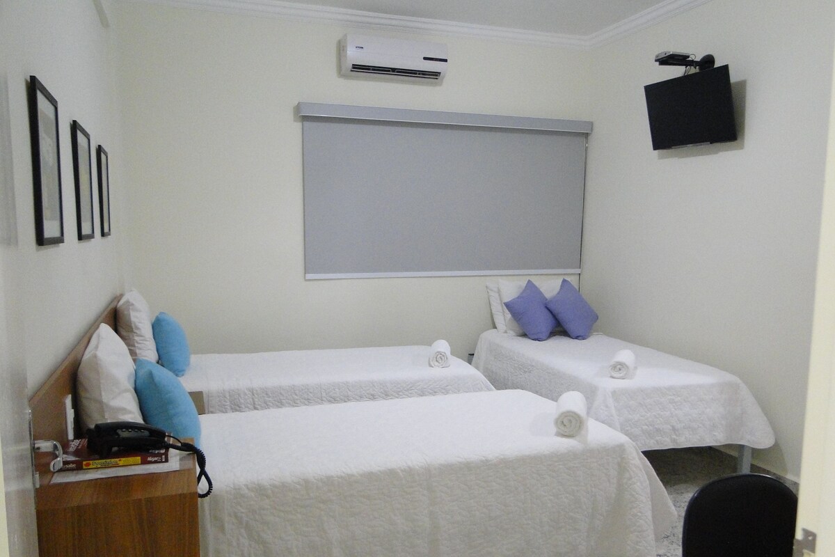A hotel room with two single beds, a desk with a telephone, a chair, a TV mounted on the wall, and an air conditioning unit.