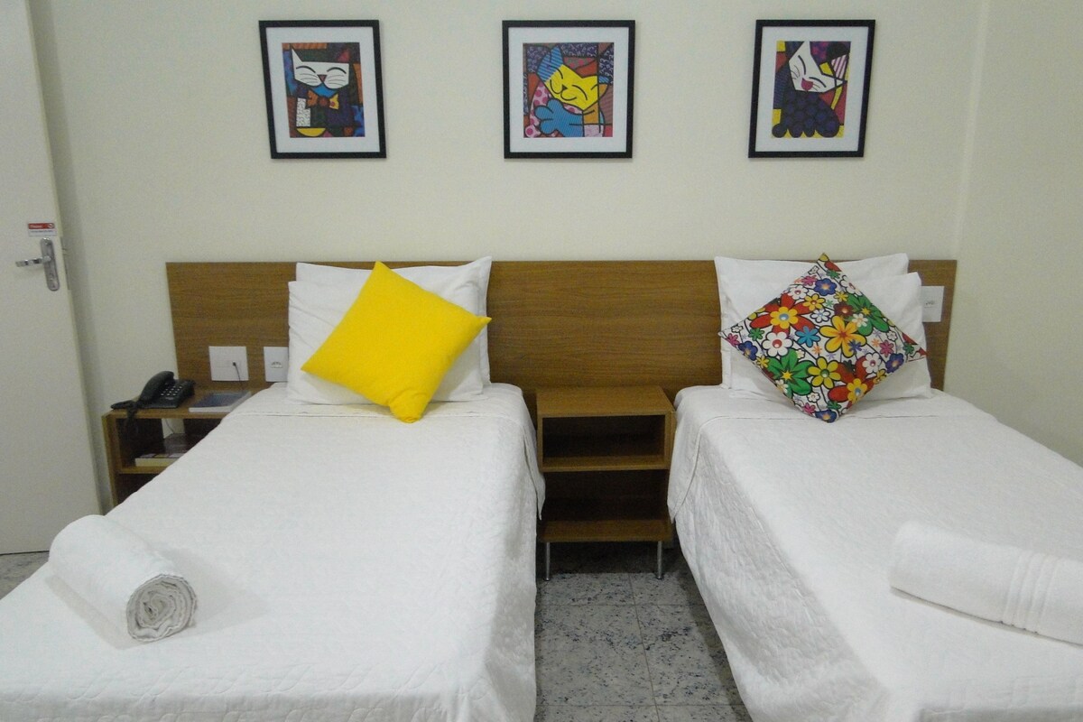 A hotel room with two single beds, a wooden headboard, a telephone on a nightstand, and three framed artworks on the wall.