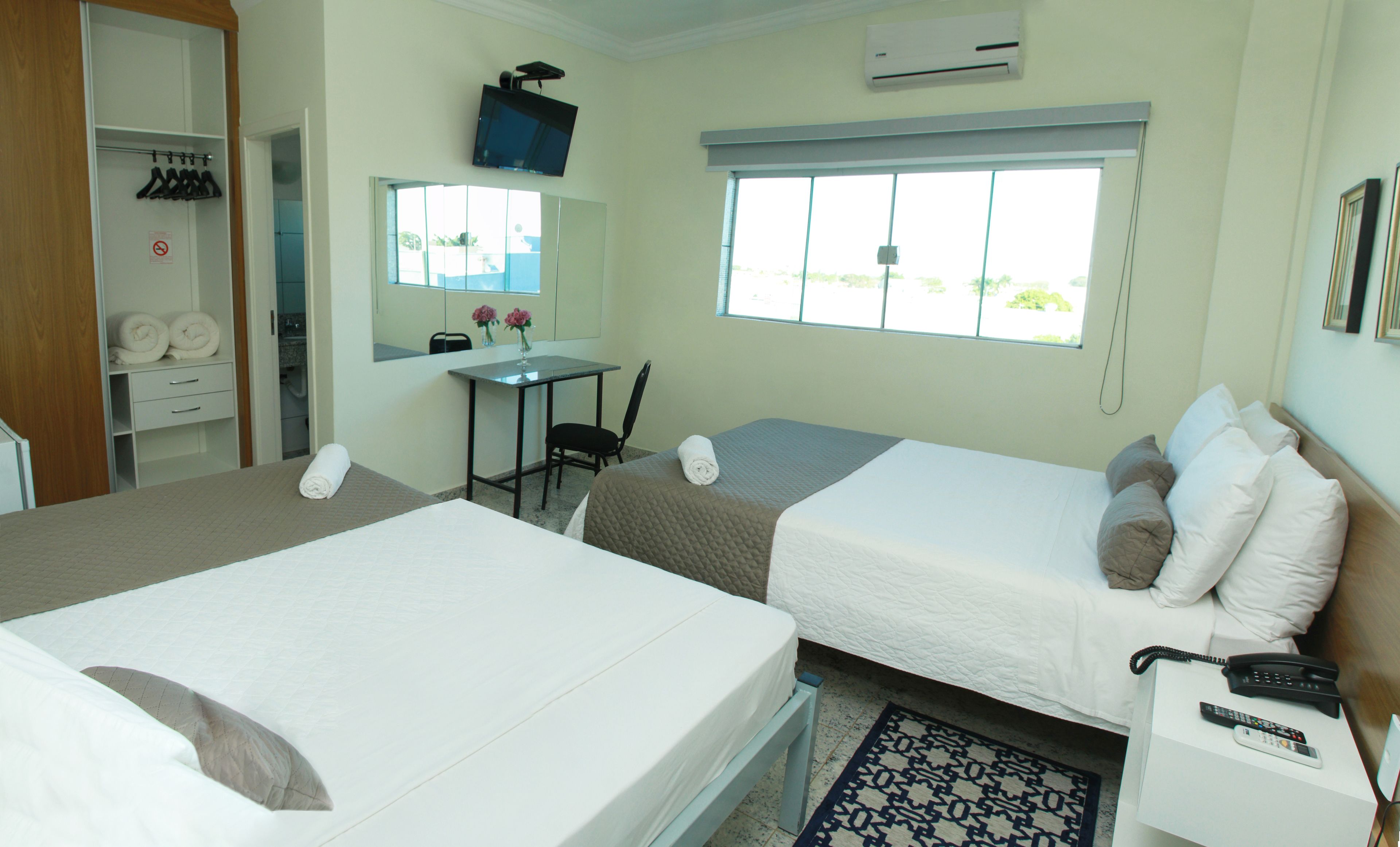 Quarto de hotel com duas camas, uma escrivaninha, uma cadeira, uma janela e banheiro.