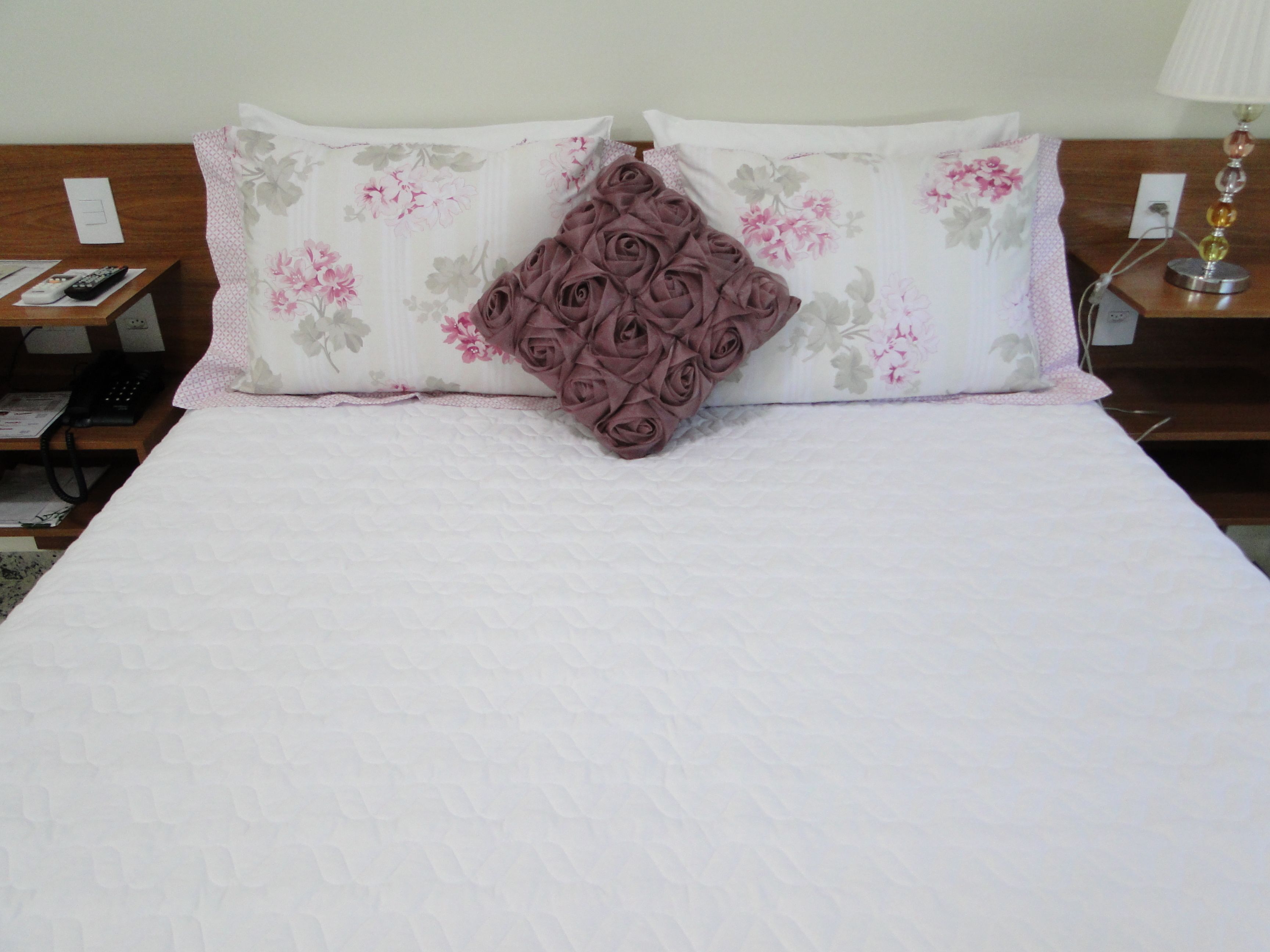 A neatly made bed with a white quilt, a floral pillow, and a decorative rose-shaped pillow.