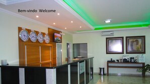 A reception area with a reception desk, multiple clocks, a television, framed artwork, and a coffee station. - Hotel Flor de Minas (Uberaba)