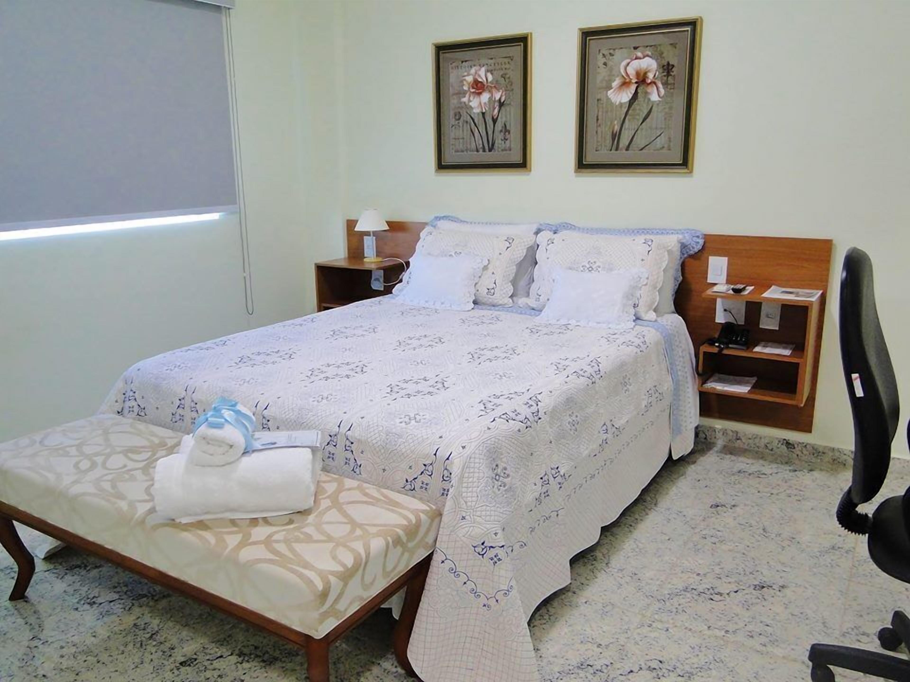 Un dormitorio ordenado con una cama, mesitas de noche, una silla y dos cuadros con motivos florales en la pared.