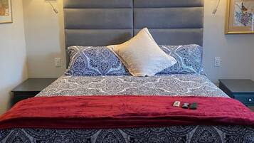 A bedroom with a patterned bedspread, a red throw blanket, and two pillows. There are two bedside tables with lamps, a wall-mounted artwork above the bed, and a framed picture on the wall.