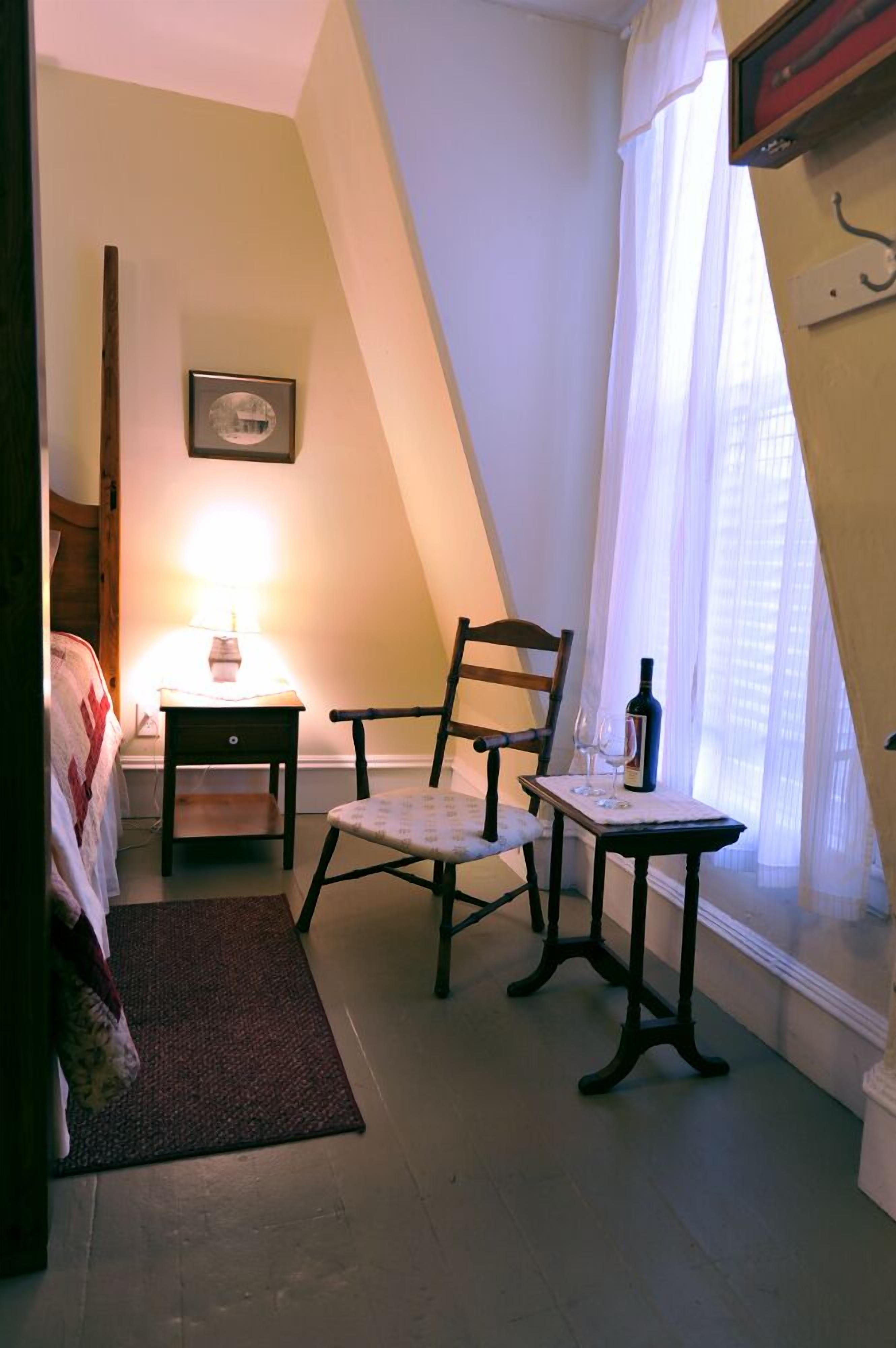 A cozy room with a bed, a chair, a small table with a bottle and glass, a lamp, and a picture on the wall.