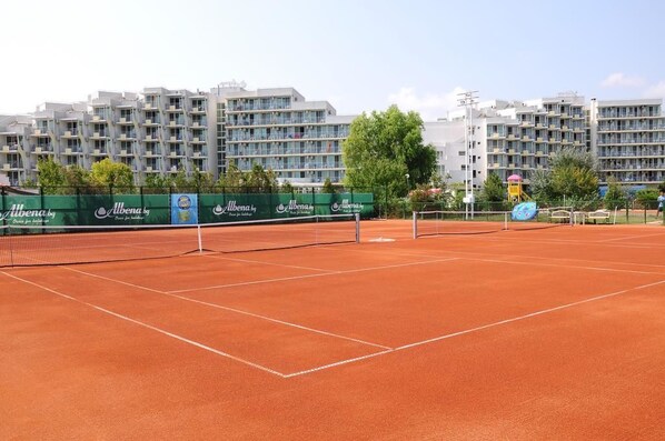 Una cancha de tenis con red, rodeada por una valla, y edificios al fondo.