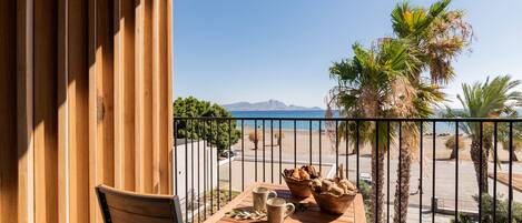 Balcón con una mesa de madera dispuesta con pan y una taza, con vista a una playa y palmeras.
