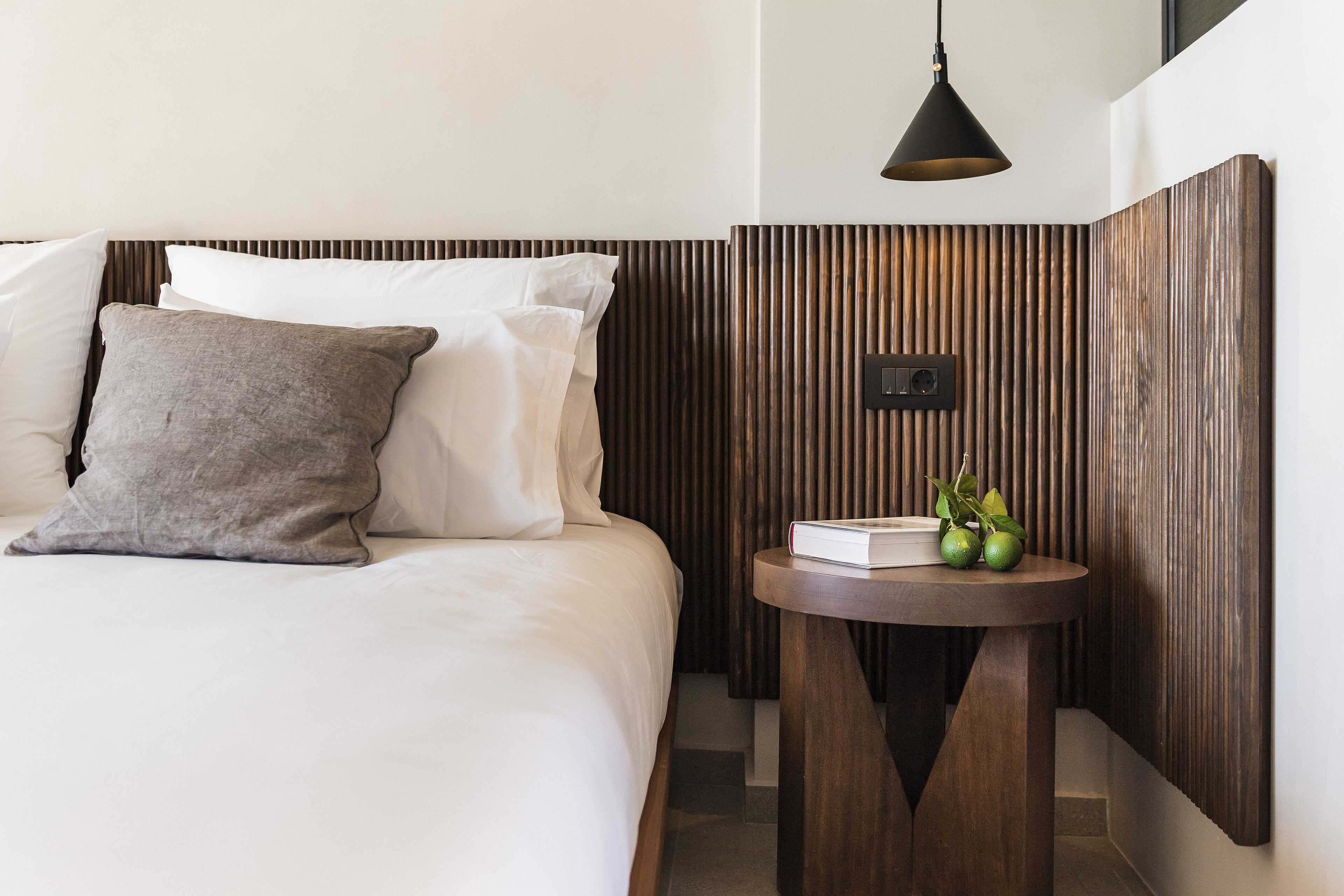 A neatly made bed with white and gray pillows, a round wooden side table with a book and two green fruits, and a modern pendant light.