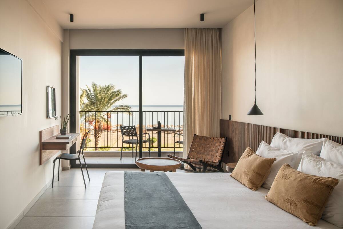 A hotel room with a large bed, a desk, a chair, a small table, and a balcony with a view of the sea and palm trees.