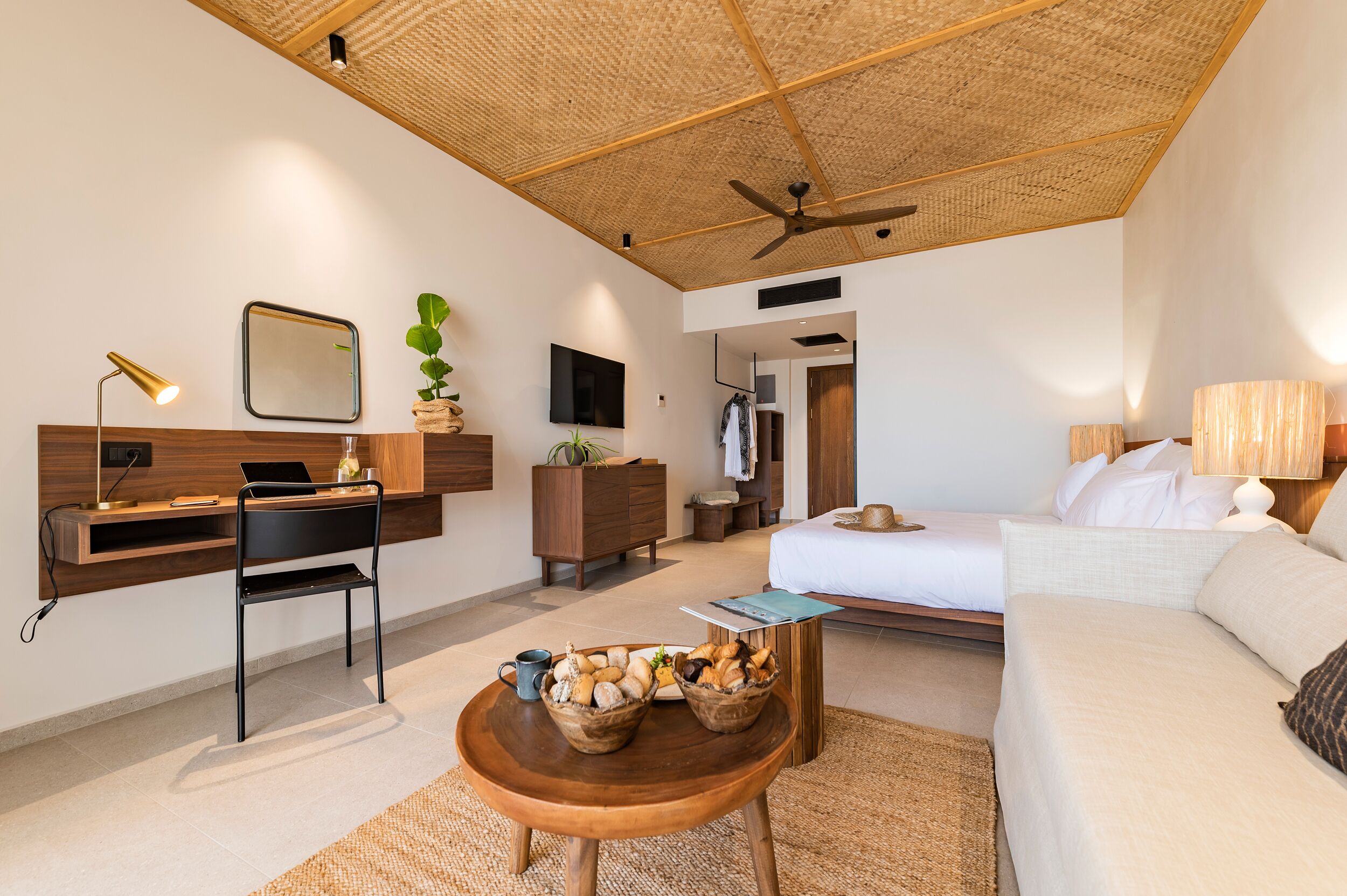 A modern hotel room with a wooden ceiling, a desk with a laptop, a bed, and a small dining table with bread baskets.