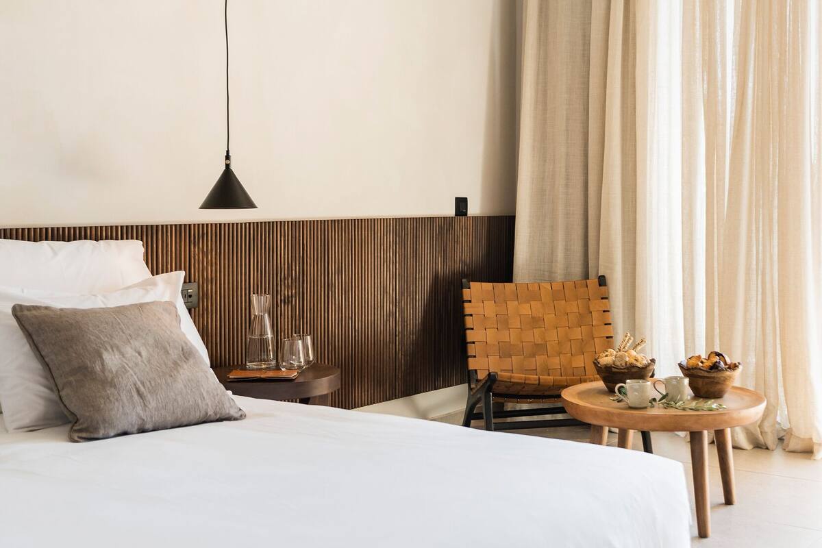 A modern hotel room with a large bed, a wooden headboard, a small table with a teapot and cups, a woven chair, and a large window with curtains.