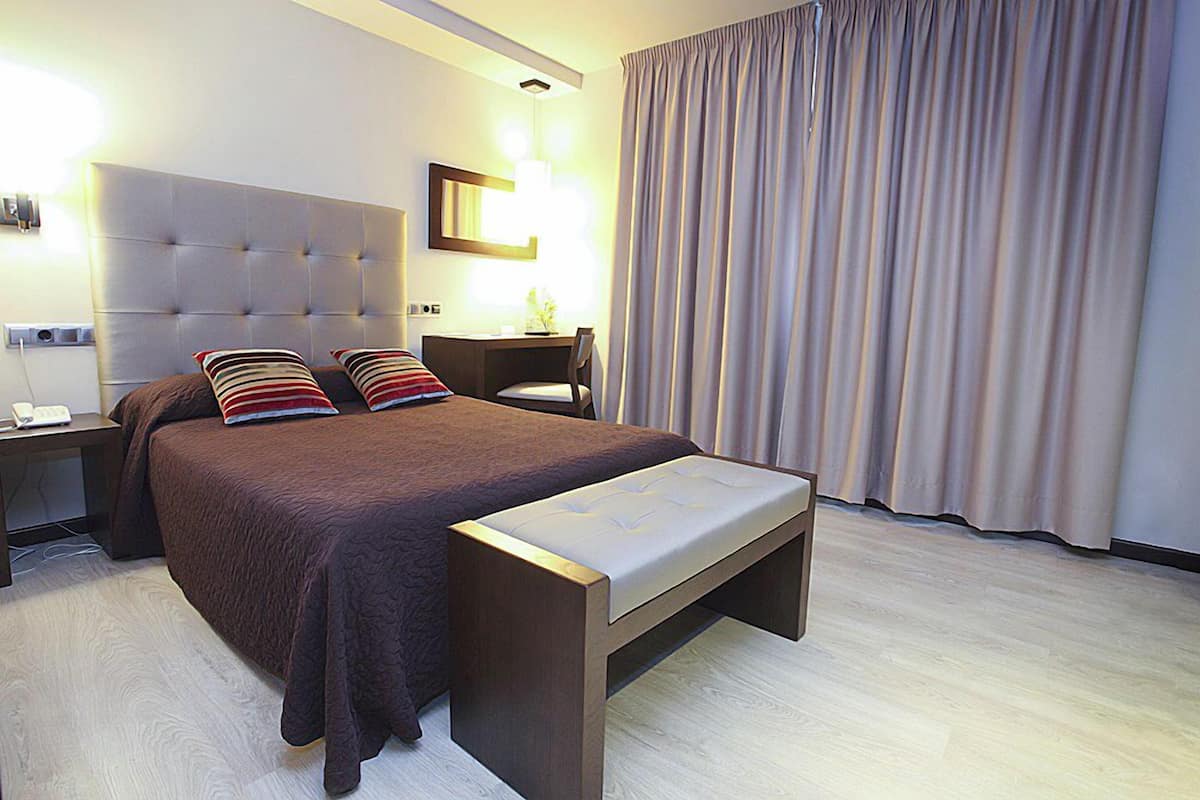 A modern hotel room with a large bed, a bedside table, a desk, and a large window with curtains.