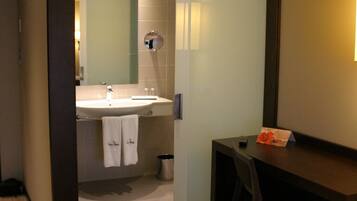 A hotel room with a wooden floor, a desk with a chair, a small trash bin, and a bathroom with a sink and towel hanging on the wall.
