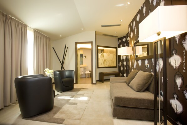 A modern living room with a sofa, a black chair, and a large black ottoman. There is a mirror on the wall and a lamp on a side table.