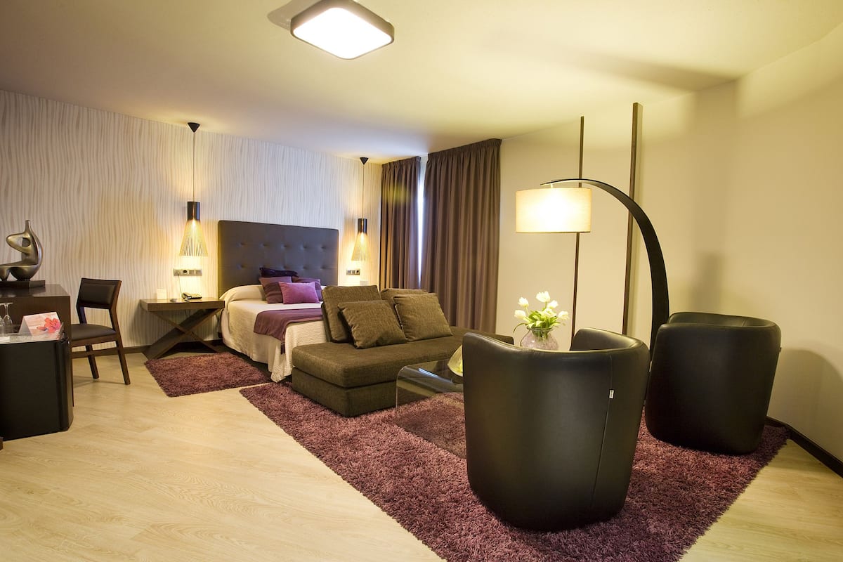 A modern hotel room with a bed, a desk, a chair, and two black ottomans.