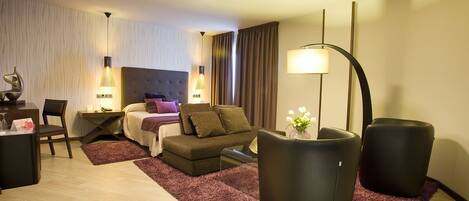 A modern hotel room with a bed, a desk, a chair, and two black ottomans.