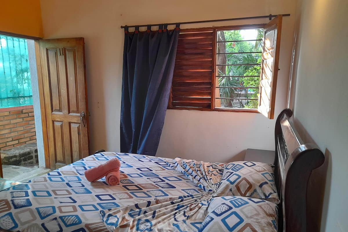 Een slaapkamer met een bed, een raam met gordijnen, een deur en uitzicht op groen buiten.