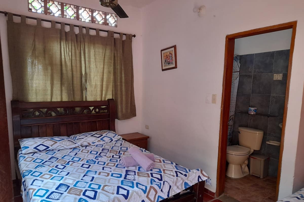 Een slaapkamer met een bed, nachtkastje, raam met gordijnen, een wandplank en uitzicht op de badkamer.