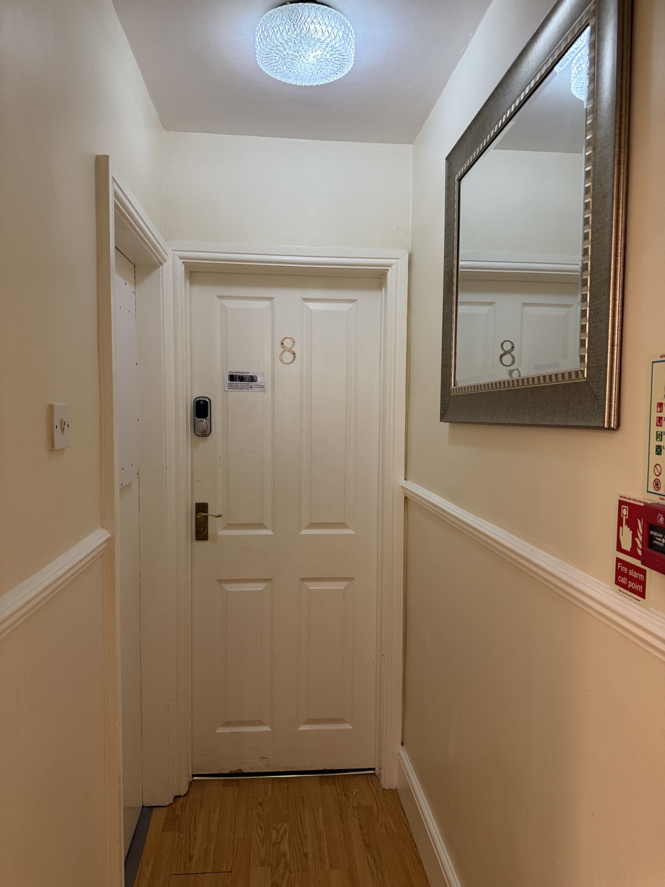 Un couloir avec une porte blanche portant le numéro 8, un miroir sur le mur et un point d’appel d’alarme incendie.