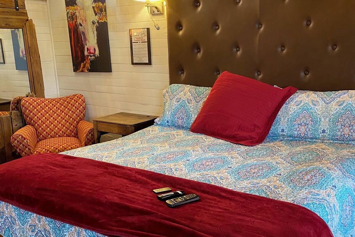 A neatly made bed with a patterned bedspread and a red throw blanket, a tufted headboard, a wall-mounted lamp, and a framed artwork.