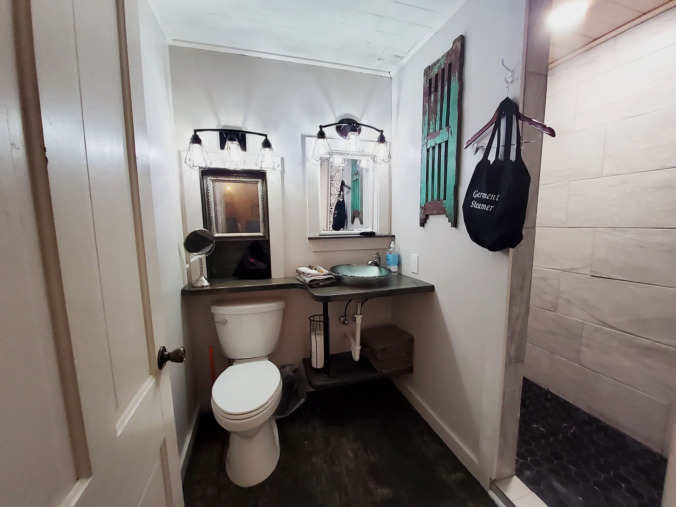 Une petite salle de bain comprenant des toilettes, un lavabo et un miroir. Un sac noir est accroché au mur.