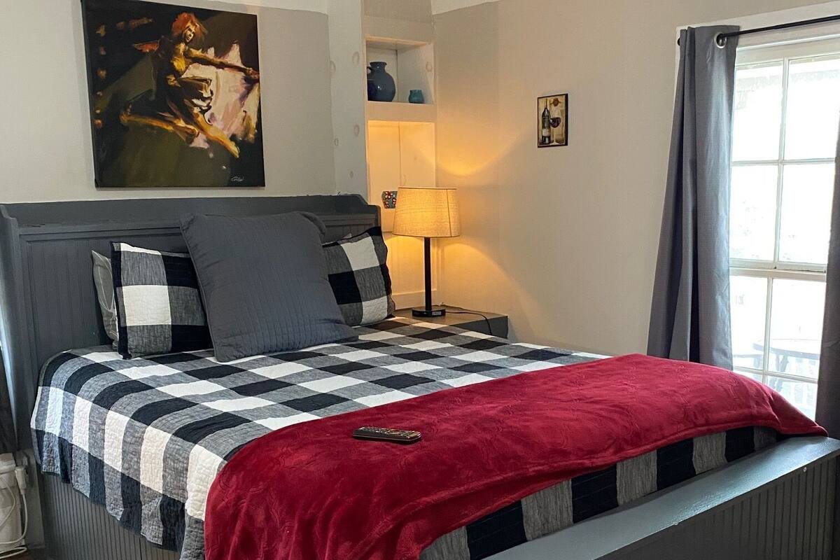 A bedroom with a bed, a nightstand, a lamp, a painting, and a window with curtains.