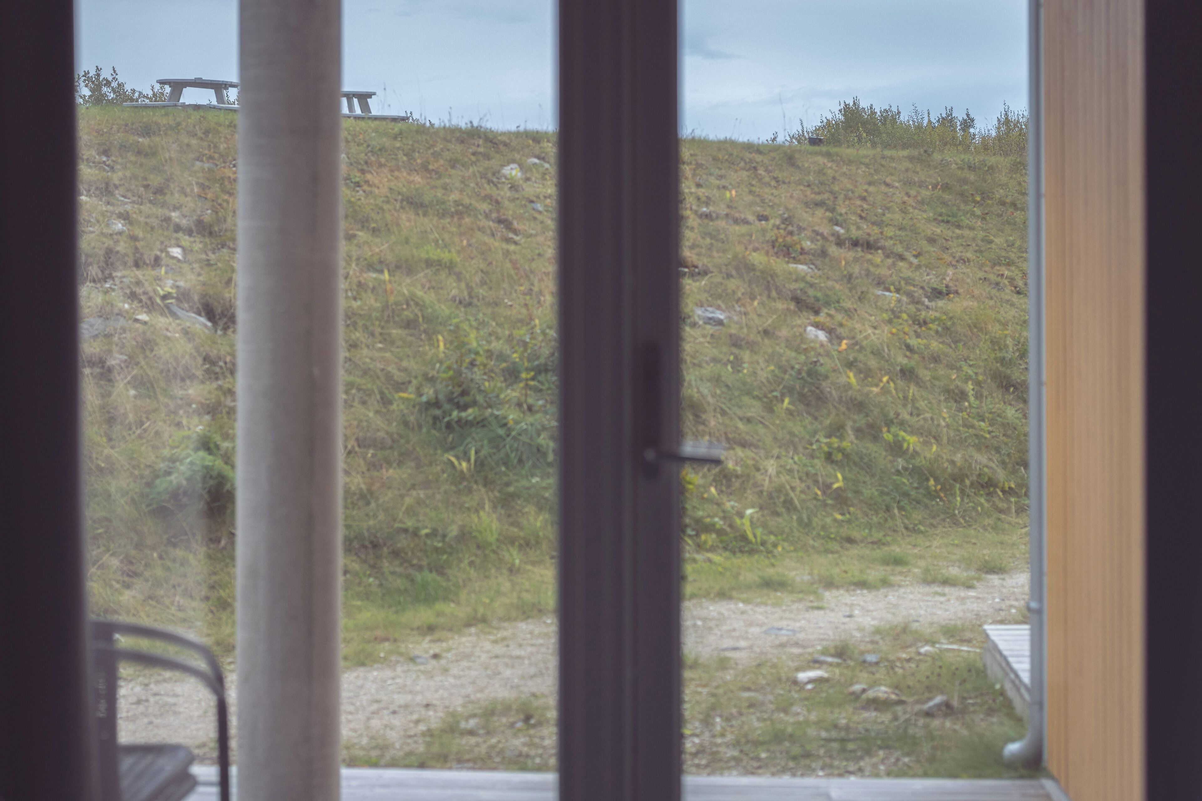A view through a window with a wooden door, a grassy hill, and a picnic table in the distance.