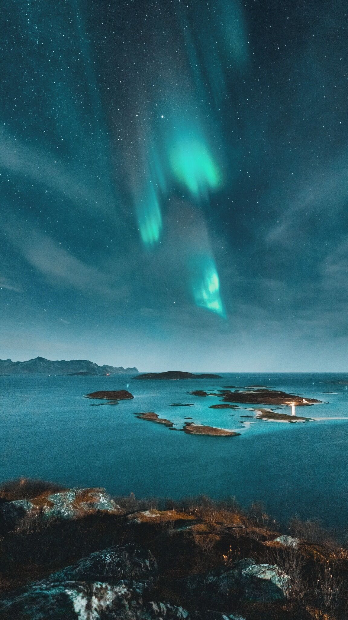 A night view of a coastal landscape with aurora borealis over a calm sea and rocky shoreline.