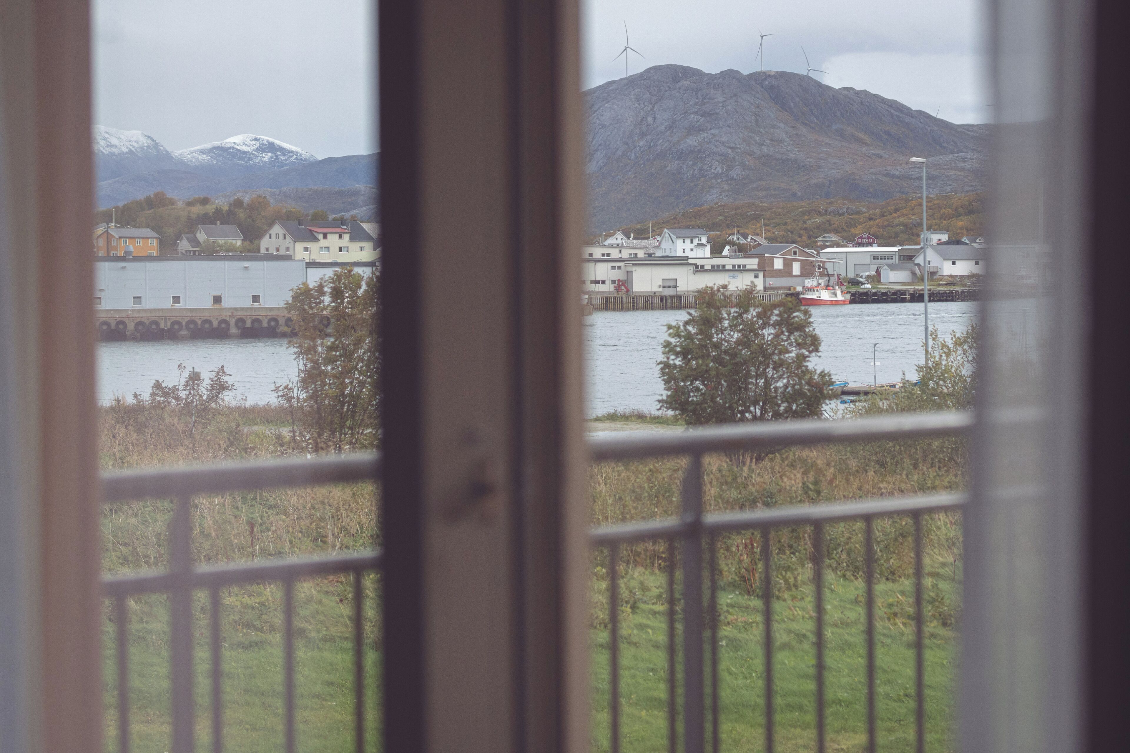 A view through a window with a metal railing, featuring a body of water, a red boat, buildings, and mountains in the background.