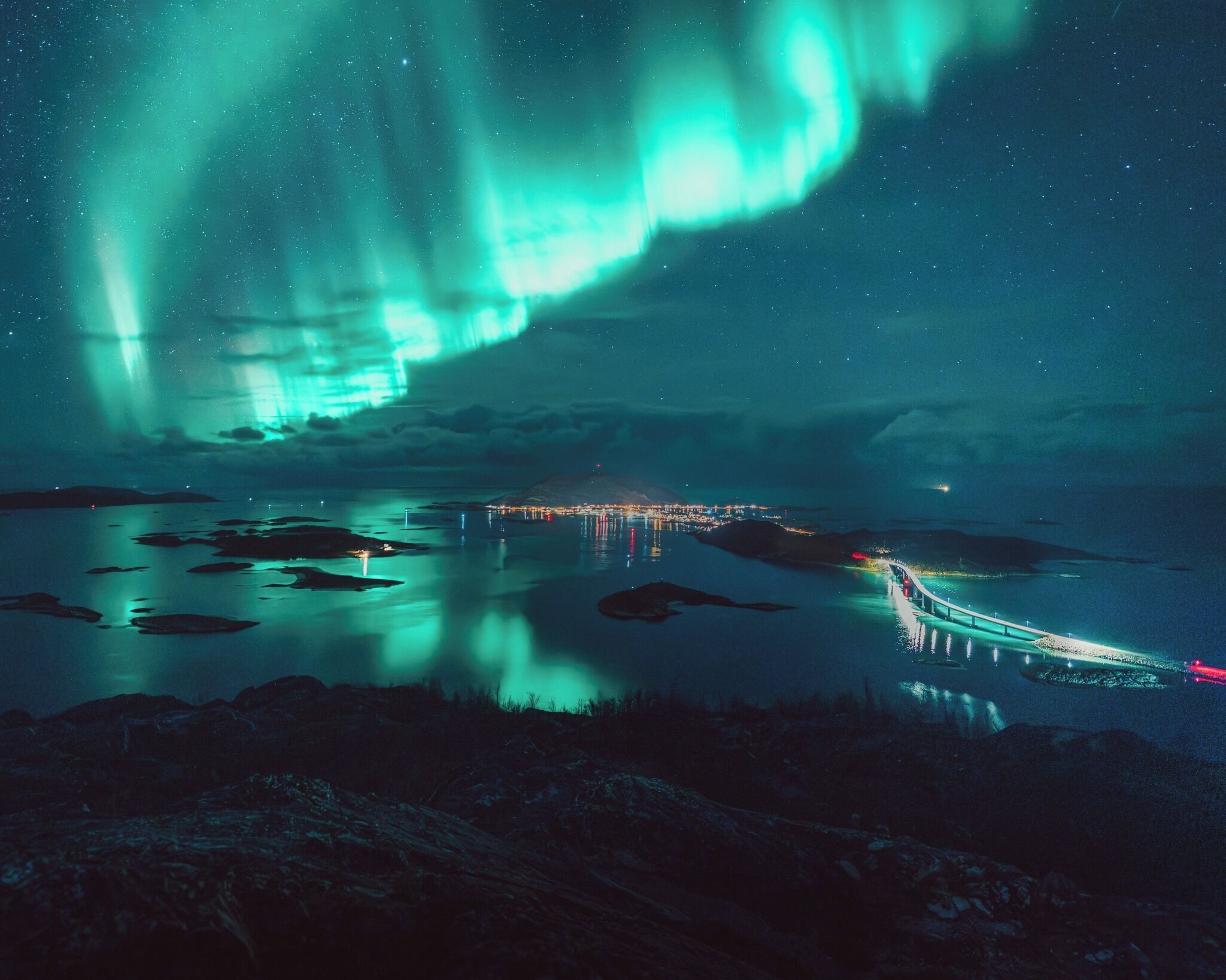 A nighttime view of a coastal area with a bridge reflecting lights, under a vibrant aurora display.