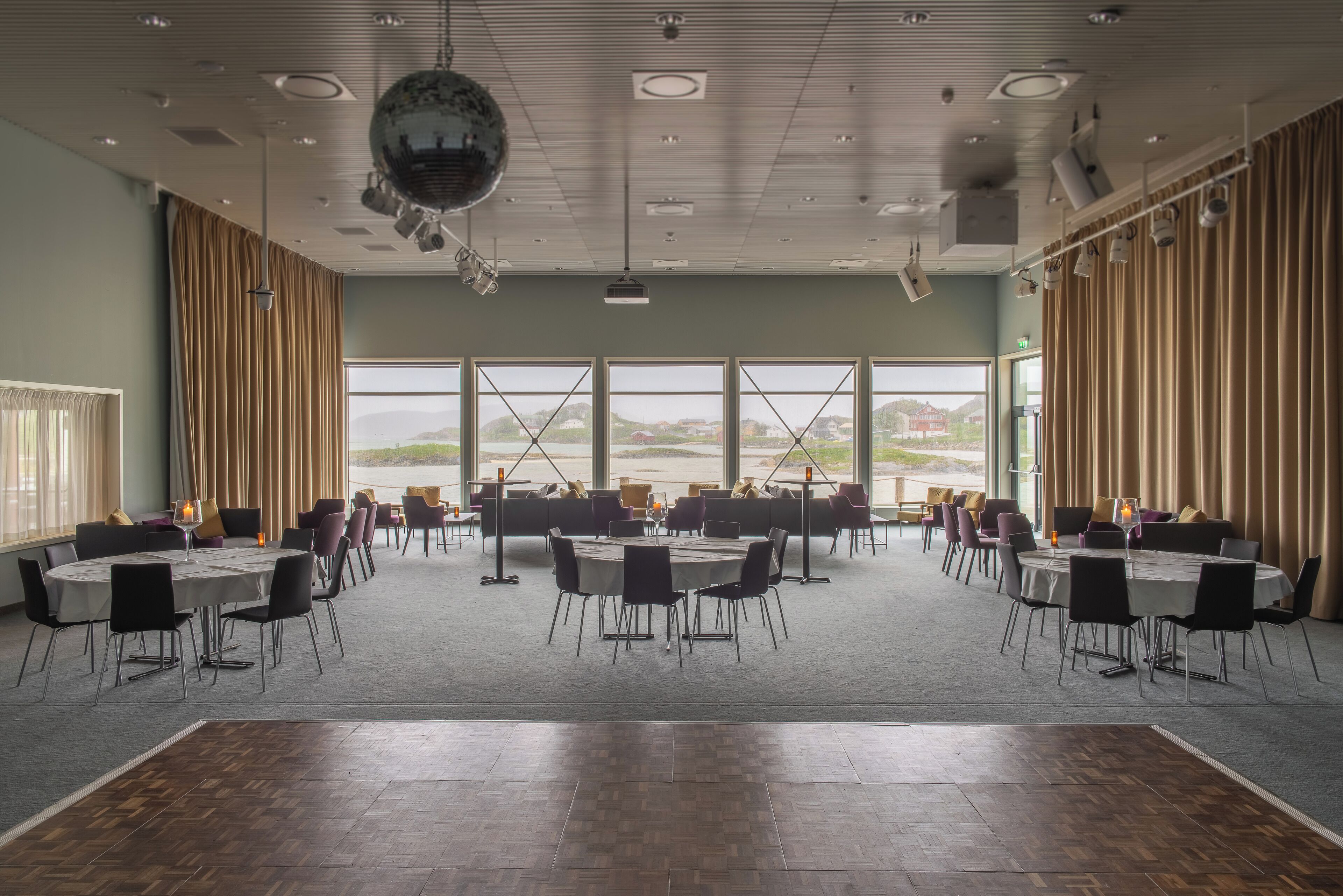 Een ruime vergaderzaal met meerdere tafels en stoelen, grote ramen met uitzicht op de omgeving, en een discobal aan het plafond.