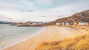 A coastal scene with a sandy beach, a row of white and yellow houses, and mountains in the background. - Sommarøy Arctic Hotel Tromsø (Tromsø)