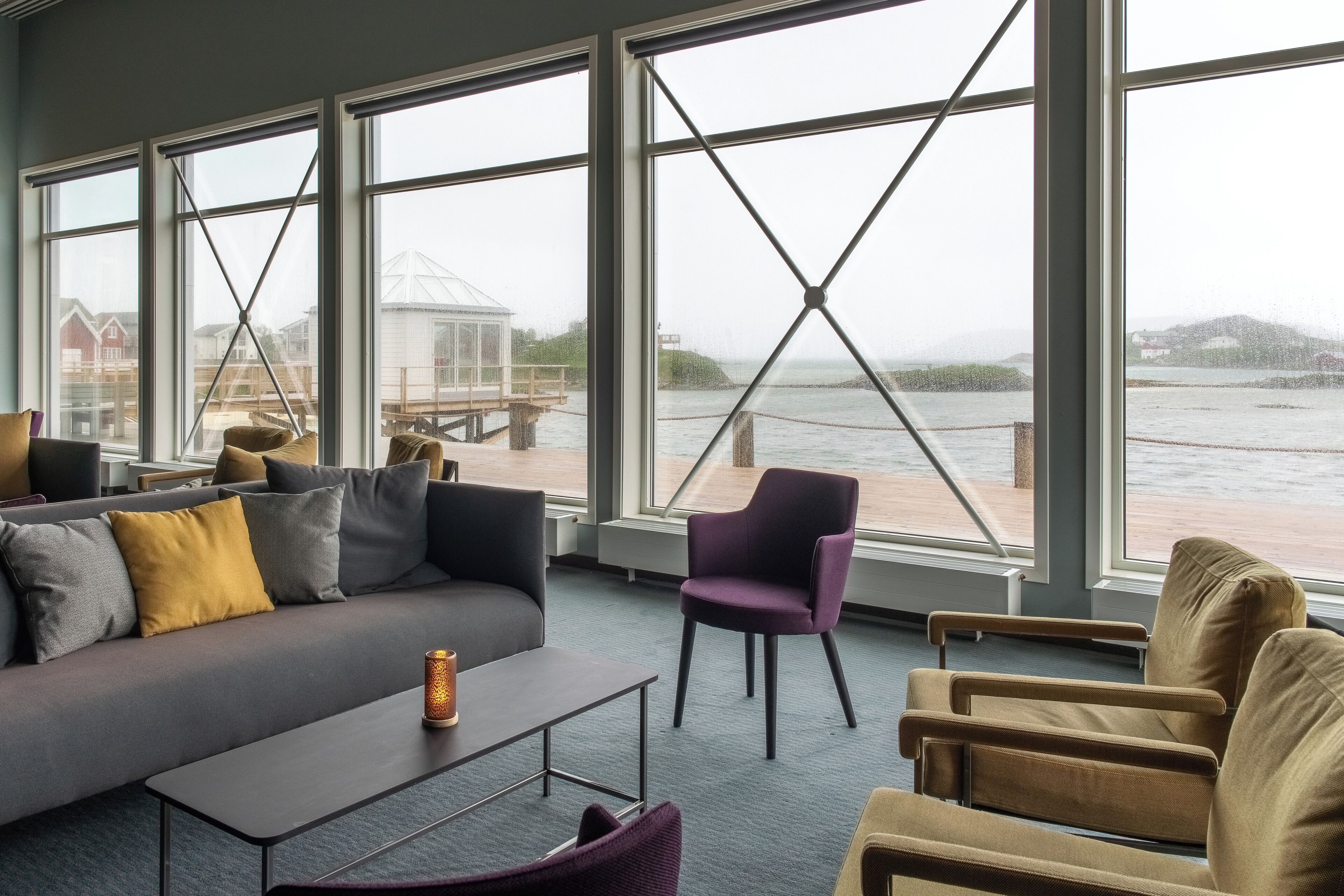 A modern living room with a grey sofa, a purple armchair, and a brown armchair. Large windows offer a view of a dock and a greenhouse.