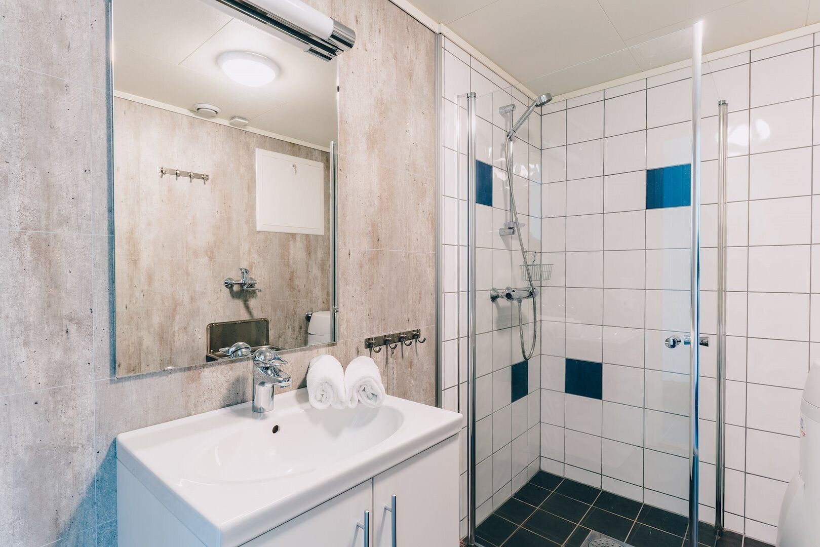 Ein modernes Badezimmer mit einem weißen Waschbecken, zwei Handtüchern, einer Duschkabine aus Glas und einem Spiegel.