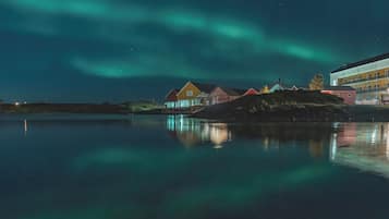 A waterfront property with colorful houses reflecting on calm water, under a night sky with the Northern Lights.