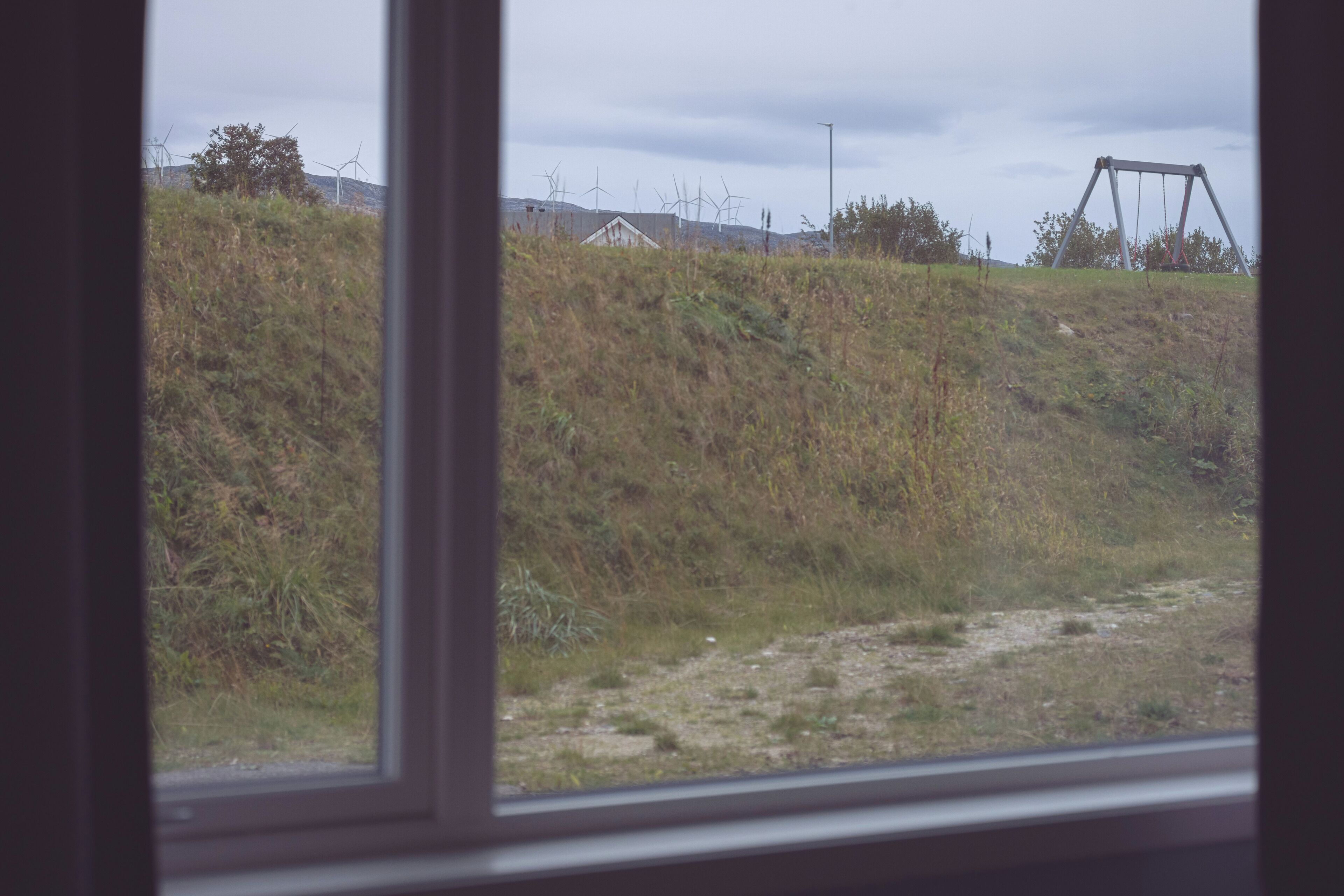 A view through a window of a grassy area with a swing set and wind turbines in the background.