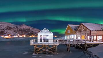 Una estructura de madera con cerramiento de vidrio, un edificio de madera con techo inclinado, y un paisaje nevado bajo un cielo nocturno con auroras boreales.