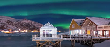 A wooden structure with a glass enclosure, a wooden building with a sloped roof, and a snow-covered landscape under a night sky with aurora borealis.